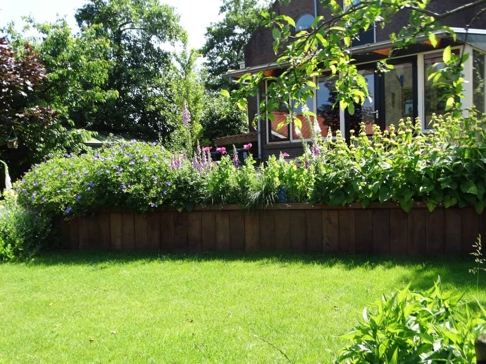 Gezicht op een tuin met bloemen, gras en terras bij een huis.