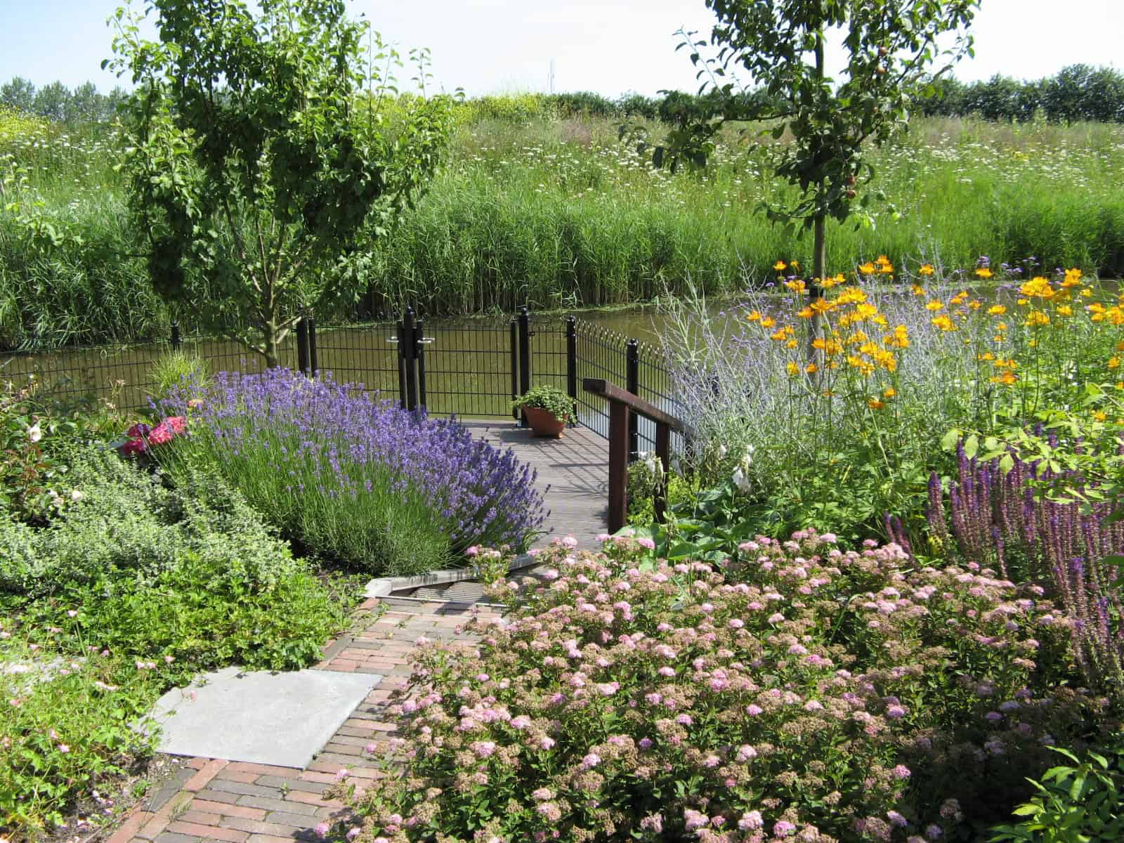 Gezicht op een bloemenrijke tuin met houten brug en waterkant.